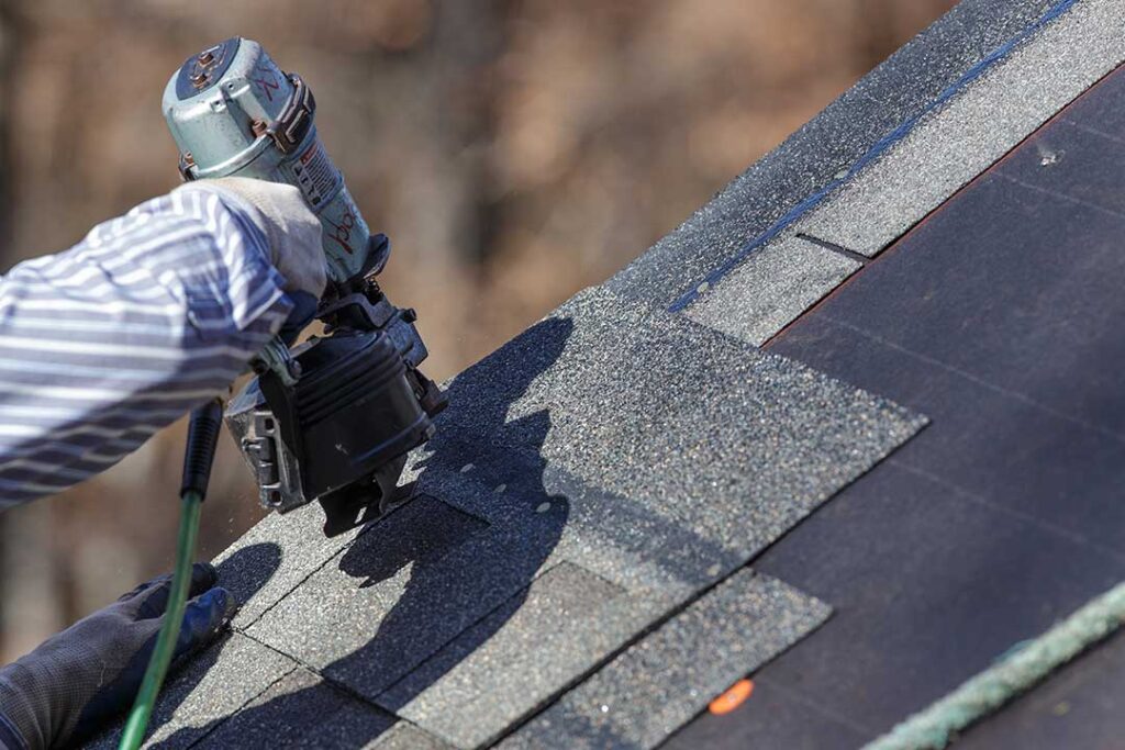 A roofer rolling out black underlayment sheets on a wooden roof deck under daylight.